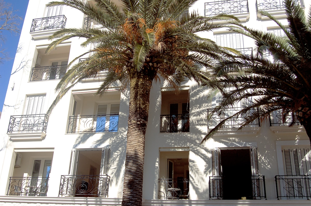 The exterior of a modern building is visible, featuring a white facade. Several balconies with ornate metal railings are seen, framed by large palm trees. The sunlight casts shadows on the building, highlighting its architectural details and inviting atmosphere.
