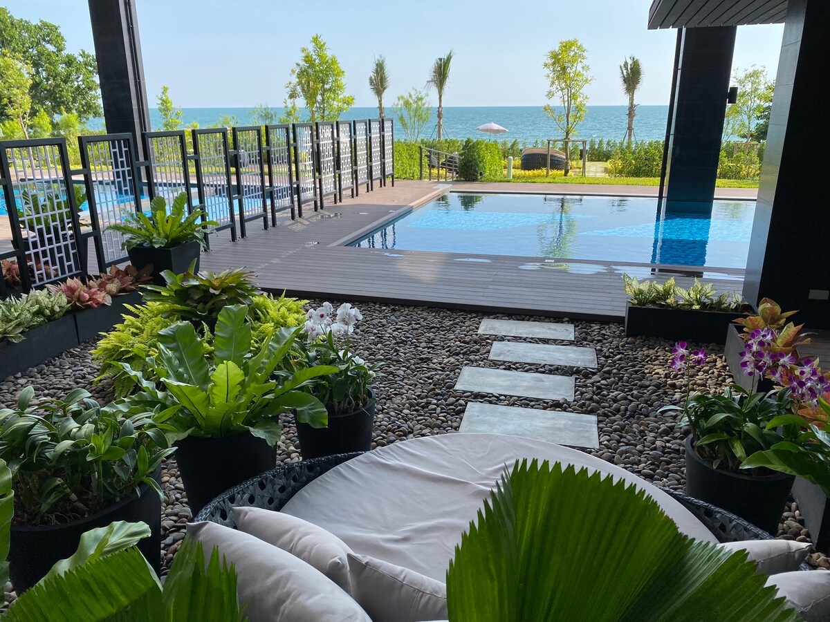 An outdoor area is featured, showcasing a serene view of the ocean and lush greenery. A spacious pool is visible alongside a path lined with stepping stones leading to a seating area adorned with various plants and flowers.