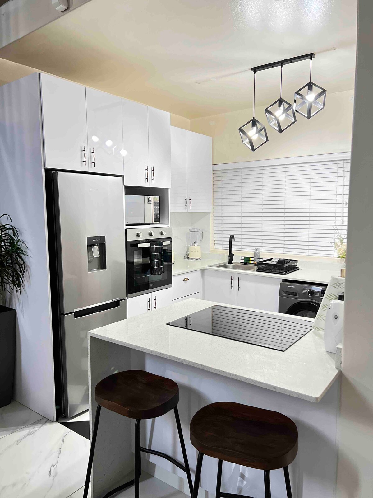 A modern kitchen features sleek white cabinetry and stainless steel appliances. A compact island offers seating for two with dark wooden stools. Natural light enters through a window adorned with blinds, and a ceiling light fixture adds contemporary elegance.