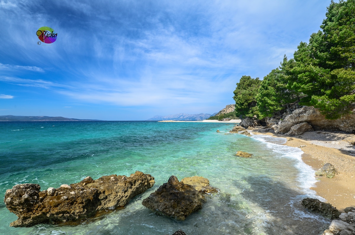 A serene beach scene is displayed, featuring clear turquoise waters lapping against a sandy shore. Naturally formed rocky outcroppings are visible along the water's edge, and lush green trees provide a backdrop under a bright blue sky with scattered clouds.