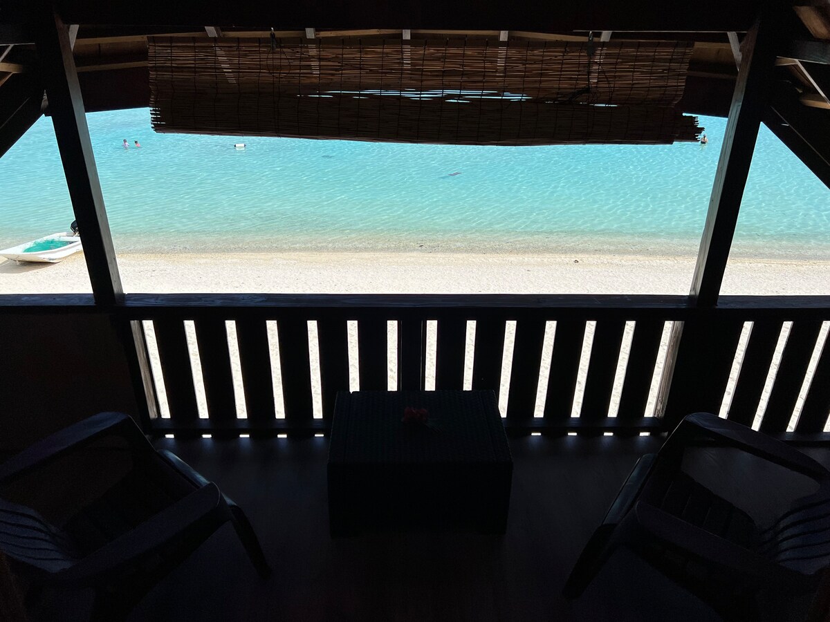 A view from a covered balcony showcases the clear turquoise waters of Matira beach. Two dark chairs are positioned on the balcony, while a small table sits in the center. The sandy beach is visible below, along with people enjoying the water.