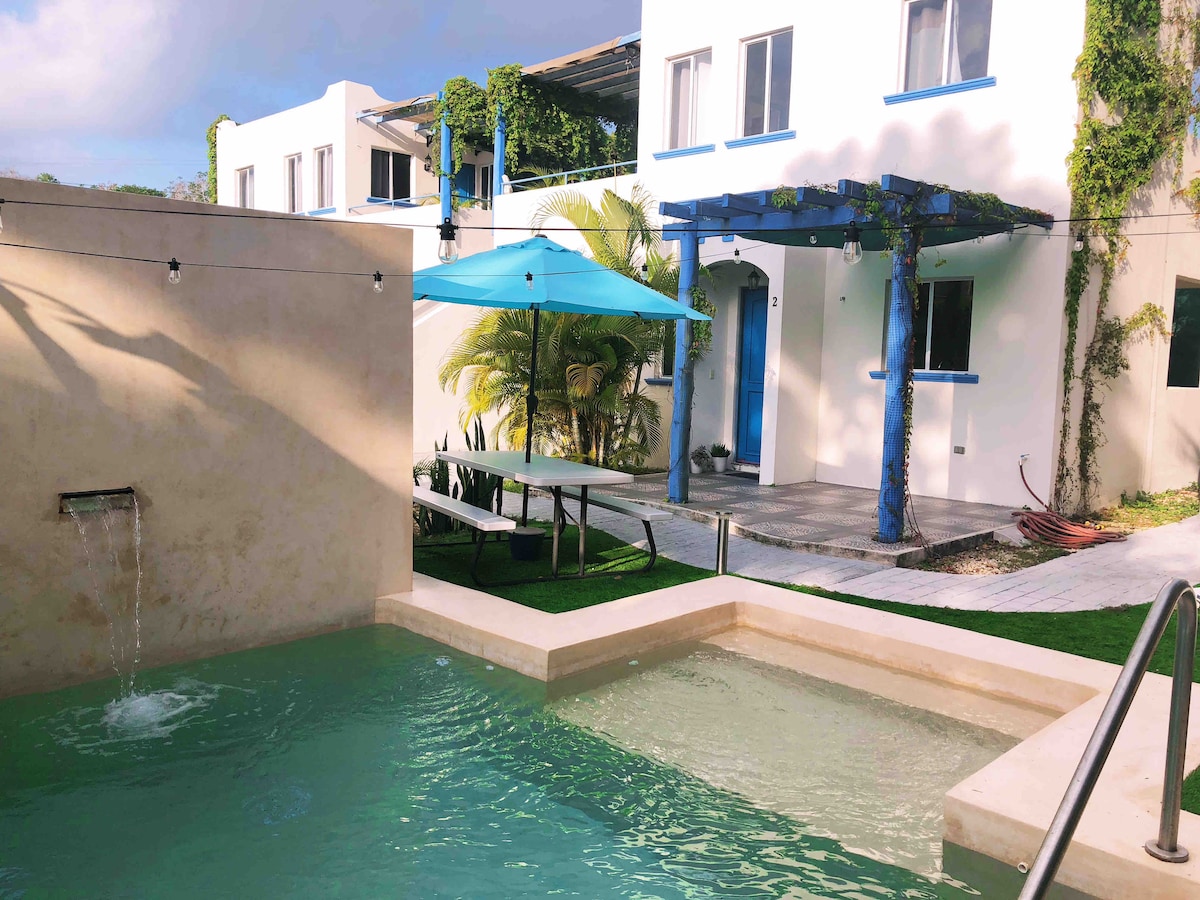 A tranquil area features a salt-water pool with gentle flowing water and a sunshade providing shade for outdoor seating. The pool is surrounded by lush greenery, with a backdrop of a two-story building adorned with vibrant blue accents and large windows, creating an inviting atmosphere.