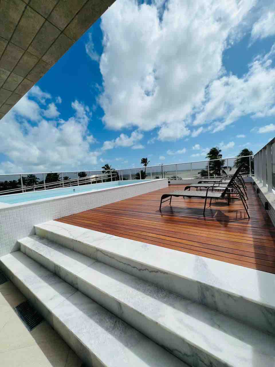A spacious wooden deck is shown, featuring several lounge chairs positioned for relaxation. Steps lead down to a pool area, with clouds scattered across a bright blue sky above. Palm trees are visible in the distance, enhancing the serene ambiance.