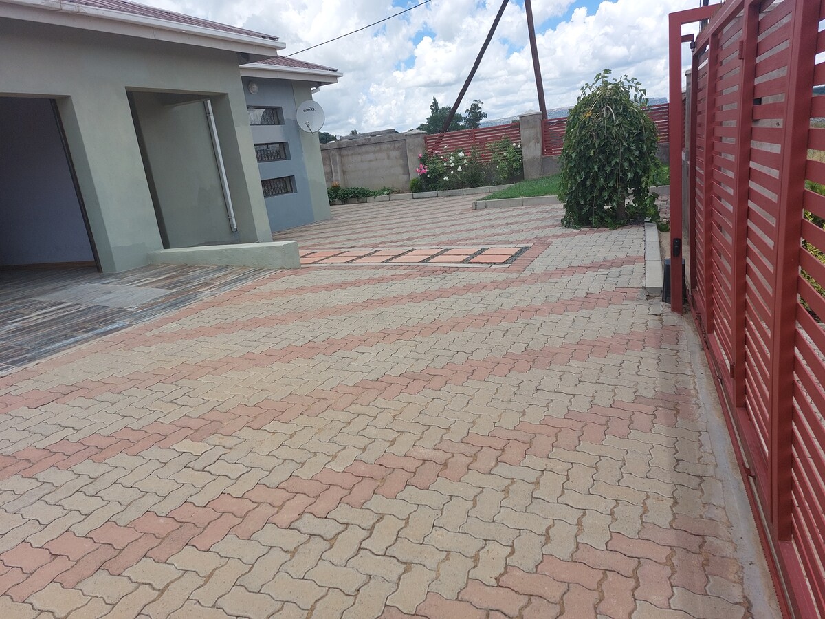 A spacious paved parking area is highlighted, featuring a combination of brick patterns in earthy tones. The area is bordered by a well-maintained garden with greenery, while a tall red gate provides secure access. The surroundings feature clear blue skies dotted with clouds.