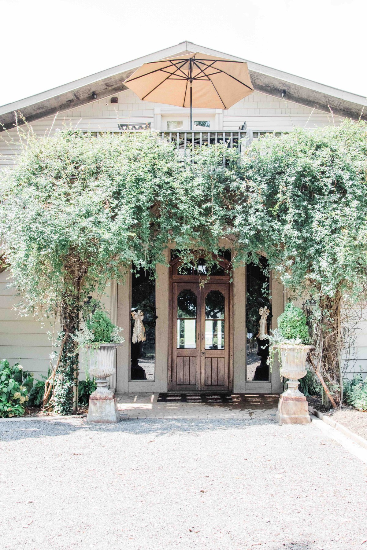 The entrance features a charming facade adorned with lush greenery, framing a set of double wooden doors. Decorative urns are placed on either side, and a large umbrella provides shade to the upper balcony, inviting visitors to appreciate the serene surroundings.