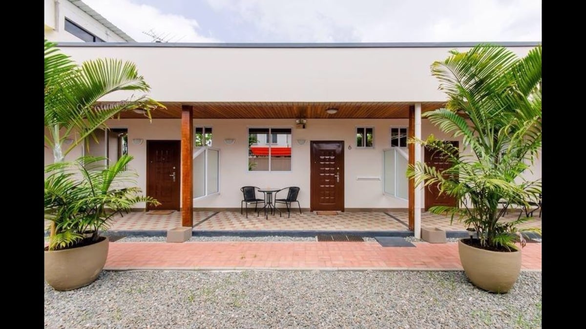 The exterior of the apartment complex features a covered walkway lined with potted palm trees. Two seating chairs and a small table are positioned on the tiled entrance. Doors to individual apartment units are framed by large windows, creating a welcoming entry area.