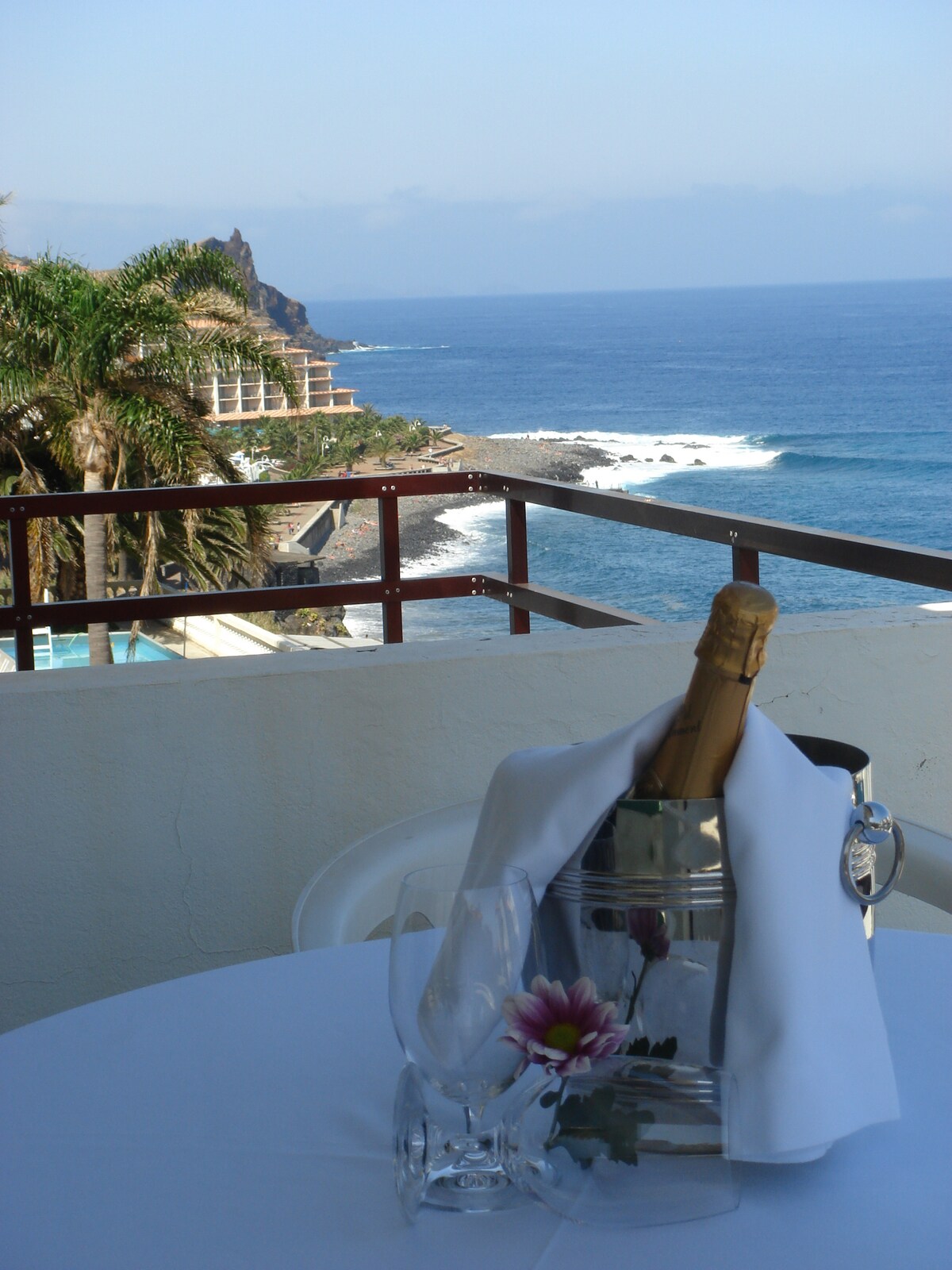 A private balcony is set with a table adorned with a flower and an ice bucket containing a bottle. The view reveals the coastline and ocean, with waves gently lapping the shore. Lush palm trees frame the scene under a clear sky.