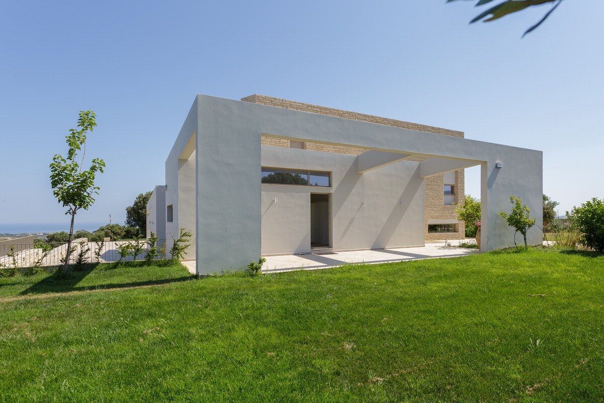 The villa exterior is displayed, featuring a modern design with clean lines and expansive windows. A manicured green lawn surrounds the structure, while the backdrop includes distant views of the sea and gentle hills. Natural light enhances the inviting atmosphere.