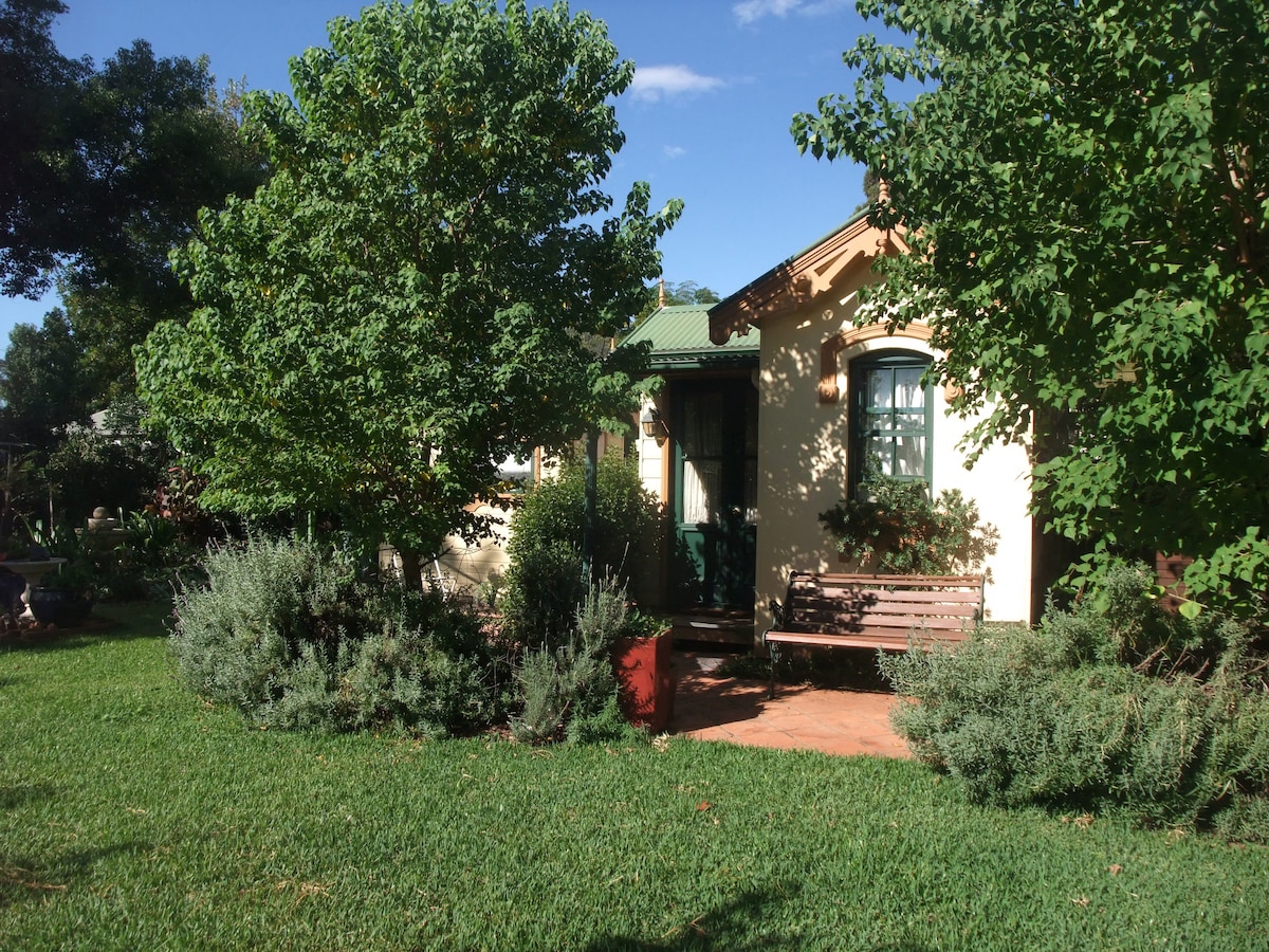A charming private room is nestled within a garden setting, surrounded by lush greenery and mature trees. A wooden bench sits invitingly on a brick path, leading to the entrance. The exterior features a warm color palette and window shutters, adding to its rustic appeal.