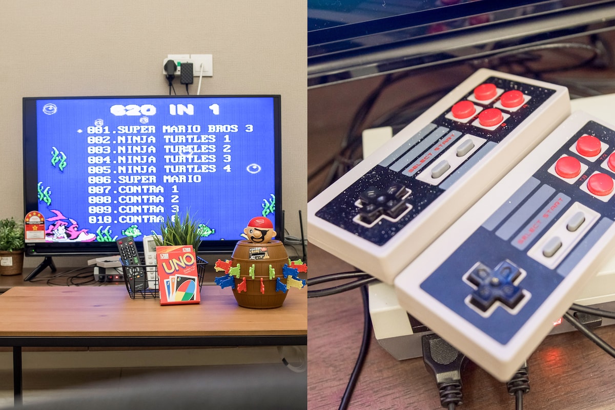 A television displays a retro gaming menu featuring various classic games, including Super Mario and Ninja Turtles. Nearby, two video game controllers are positioned on a desk, showcasing multiple buttons and a nostalgic design. The setting is completed by decorative items, including a colorful plant and a board game.
