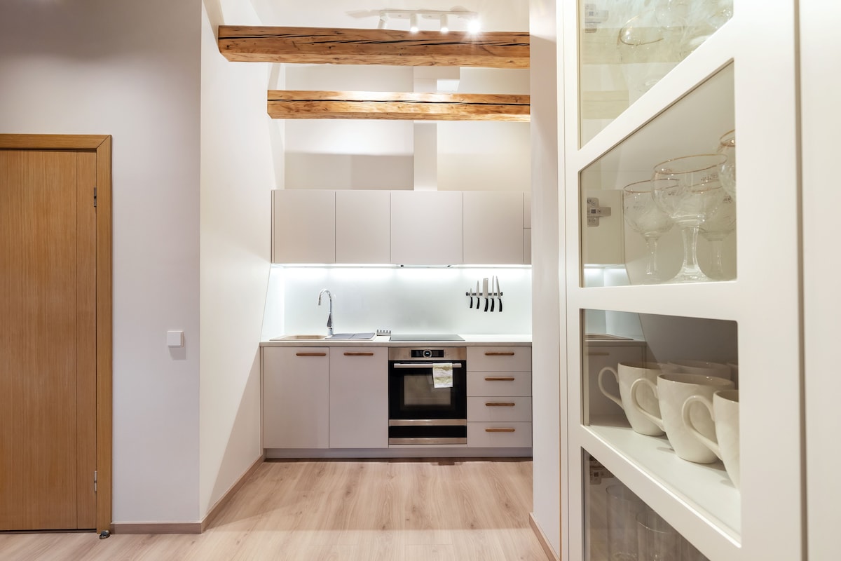 A modern kitchen is displayed with sleek white cabinetry and a minimalist design. It features high-end appliances, including an oven with a microwave. Wooden beams run across the ceiling, adding warmth to the space, while glassware and crockery are neatly arranged in a nearby cabinet.