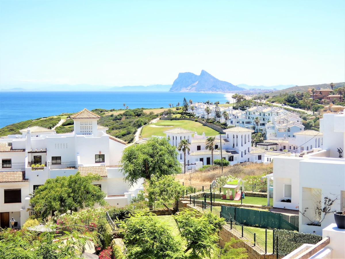A panoramic view showcases the coastline with the Mediterranean Sea and a distant view of Gibraltar. Lush greenery and white buildings are visible in the foreground, and the golf course meanders through the landscape. Clear blue skies enhance the vibrant colors of the scene.