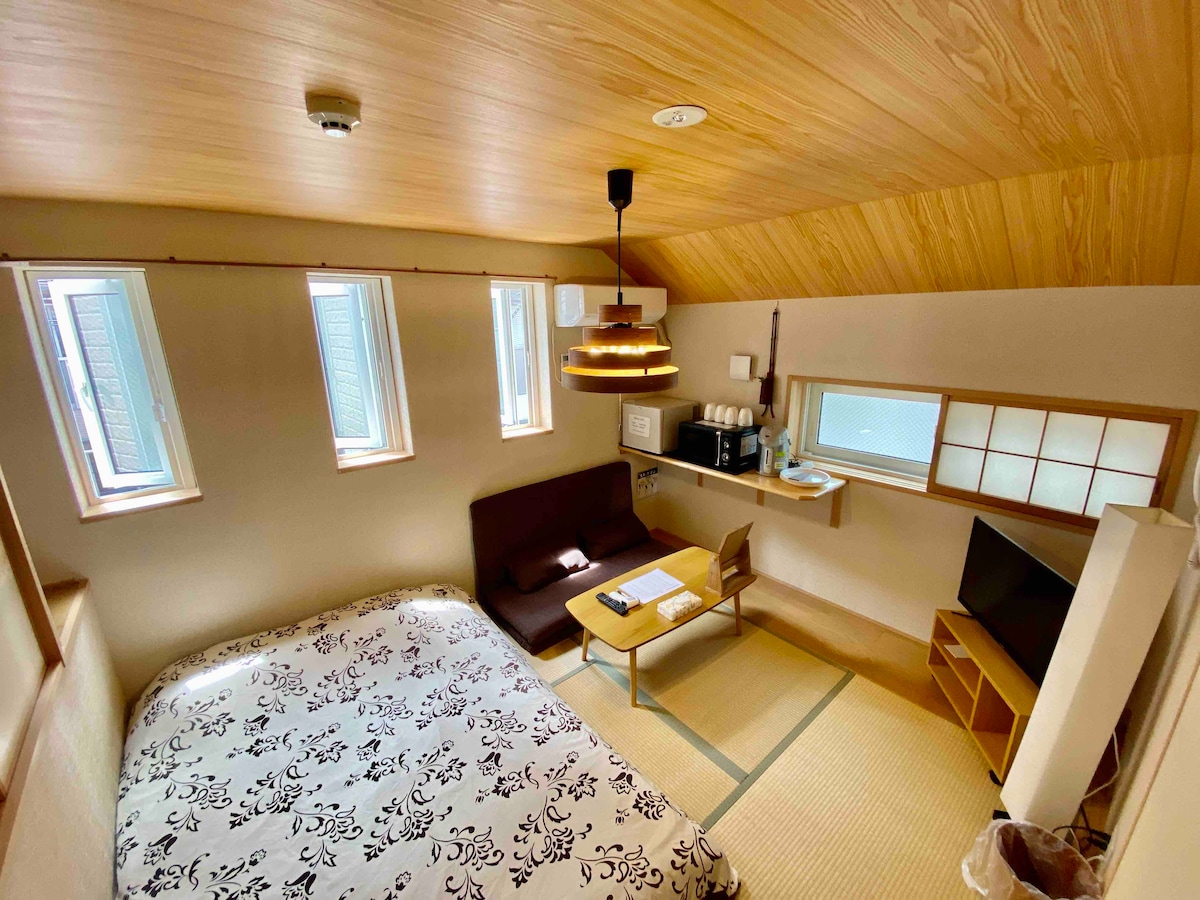 A room featuring a spacious bed with patterned bedding, a brown sofa, and a small dining table. Light streams in through multiple windows, illuminating the light wood ceiling. A kitchenette is visible, with a television near the entrance.