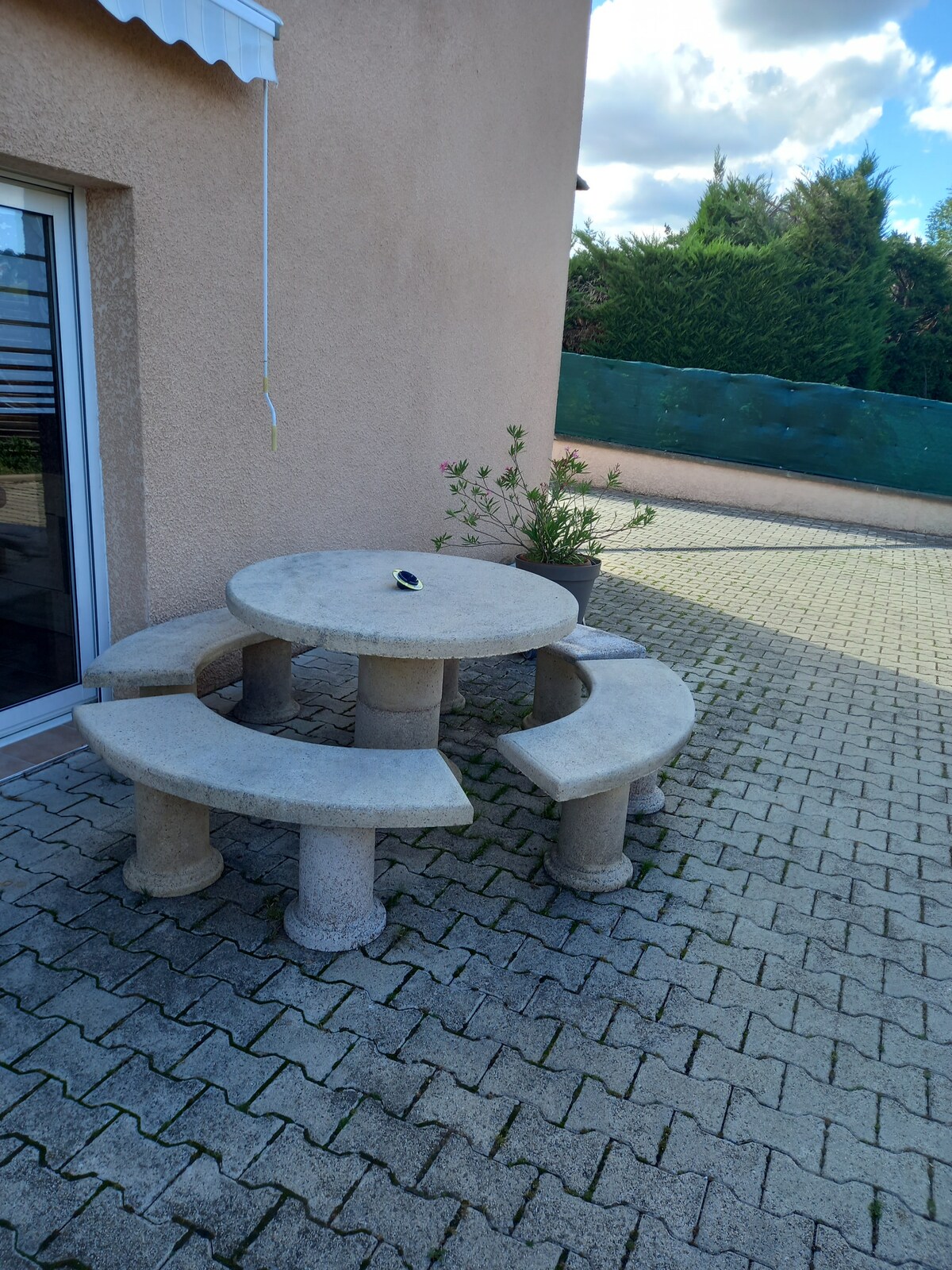 A stone dining table is surrounded by three curved benches, situated on a paved terrace. Greenery is present in a potted plant nearby, with a soft light illuminating the area from above. The exterior wall of the house forms the background.