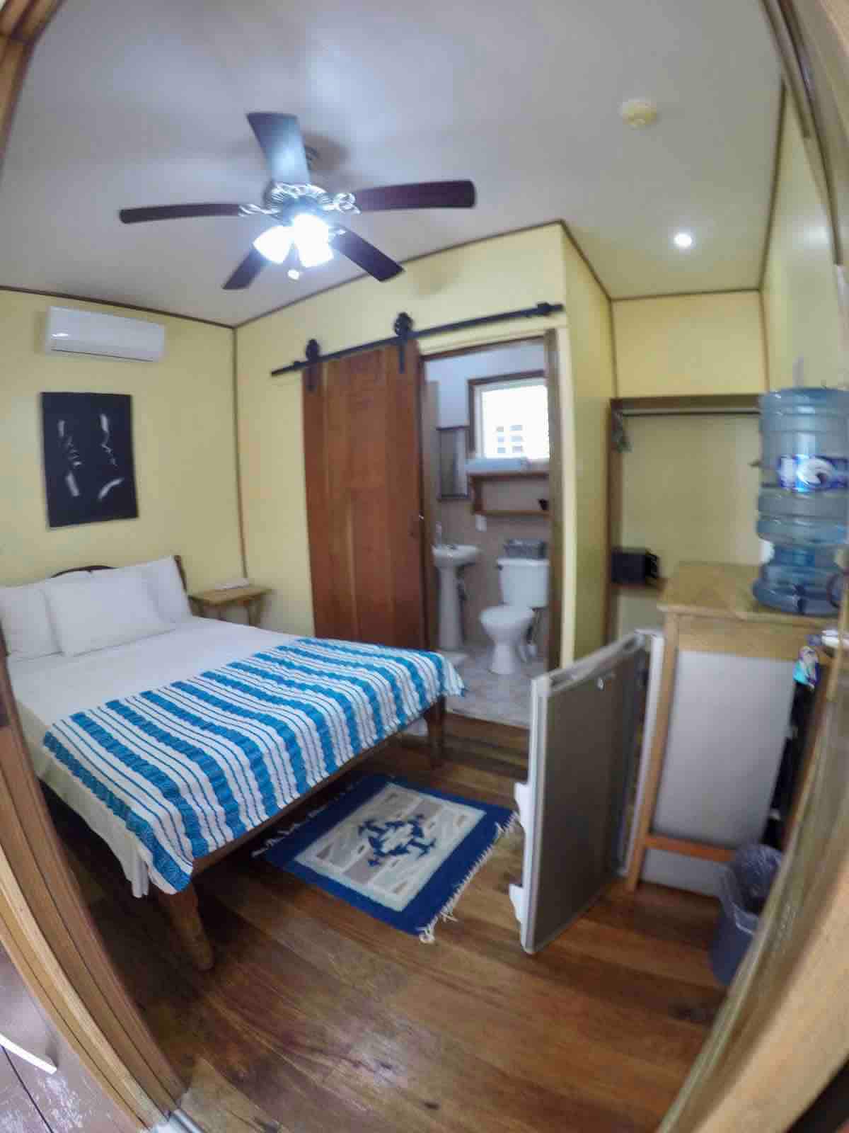 A cozy room is depicted, featuring a spacious bed with a striped blue and white blanket. A ceiling fan is visible above, and a bathroom is accessed through a door. Additional details include a small wooden table and a water cooler in the corner.