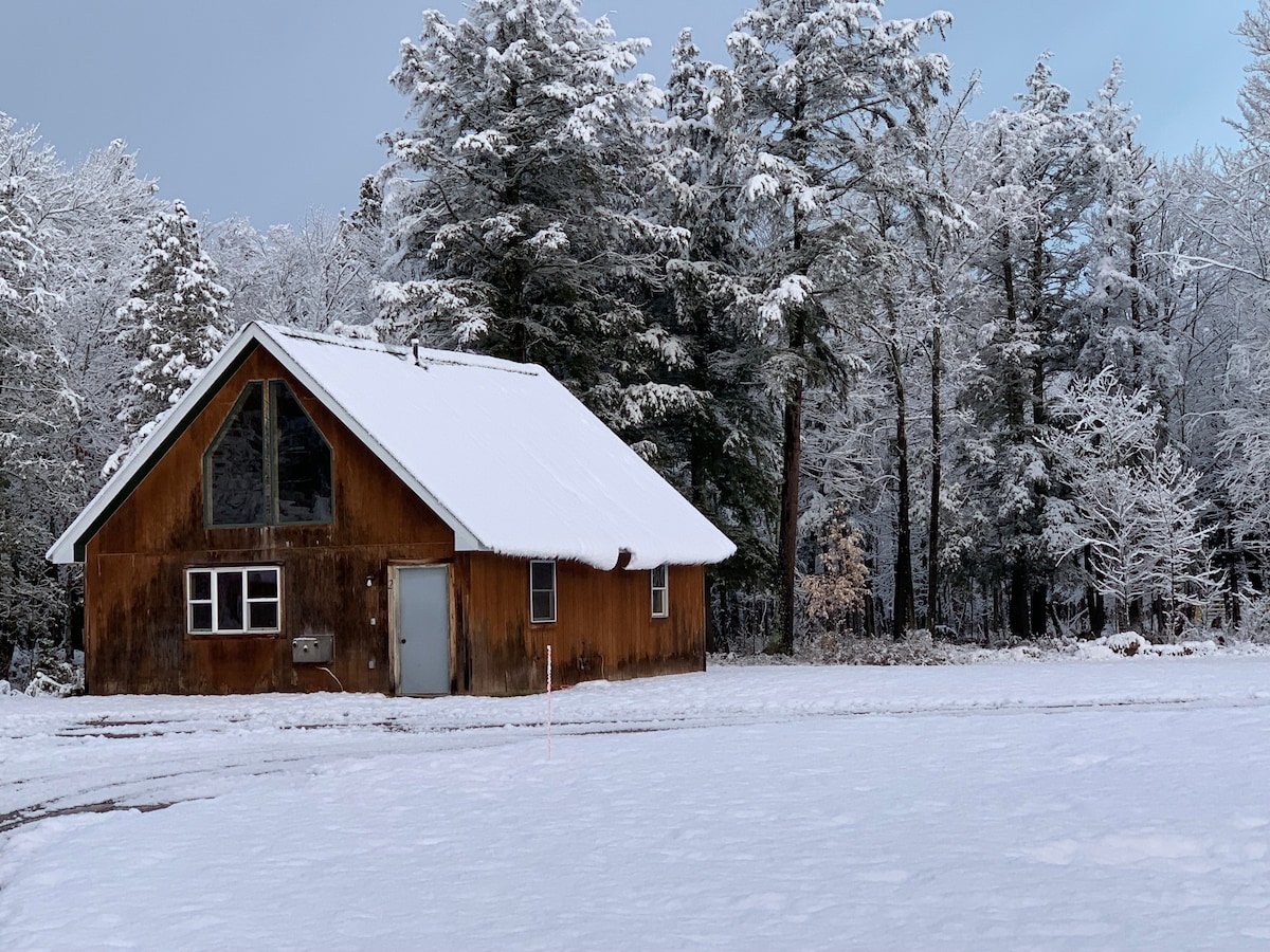 A wooden cabin is shown amidst a blanket of fresh snow, surrounded by tall pine trees. The sloped roof is dusted in white, while a large window reflects the snowy landscape. The path leading to the entrance is visible, framed by untouched snow.