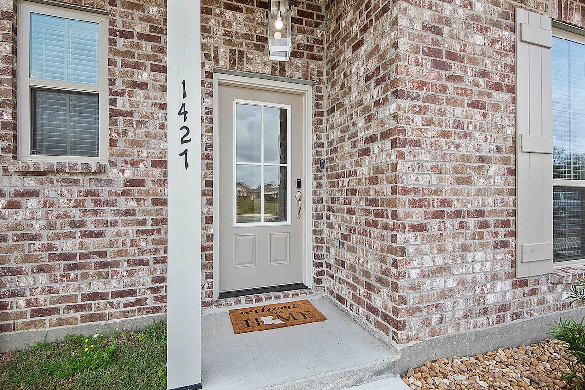 The welcoming entrance features a brick façade with large windows allowing natural light to filter in. A keyless entry door with a simple design is complemented by a doormat that reads 'welcome HOME,' encouraging a sense of arrival.