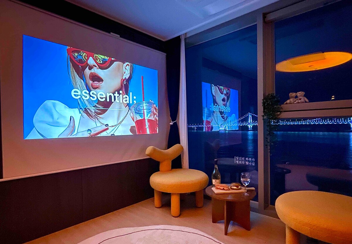 A cozy living area features a wall-mounted projection screen displaying a colorful image. A round table is set with two glasses and a bottle, while a unique yellow chair provides seating. Large windows showcase a view of the bridge and water outside.
