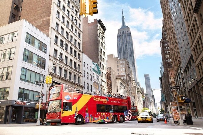 A busy city street scene is captured, featuring a bright red double-decker bus with colorful graphics. Tall buildings line the street, with the iconic Empire State Building visible in the background under a clear sky. Taxis and pedestrians add to the urban atmosphere.