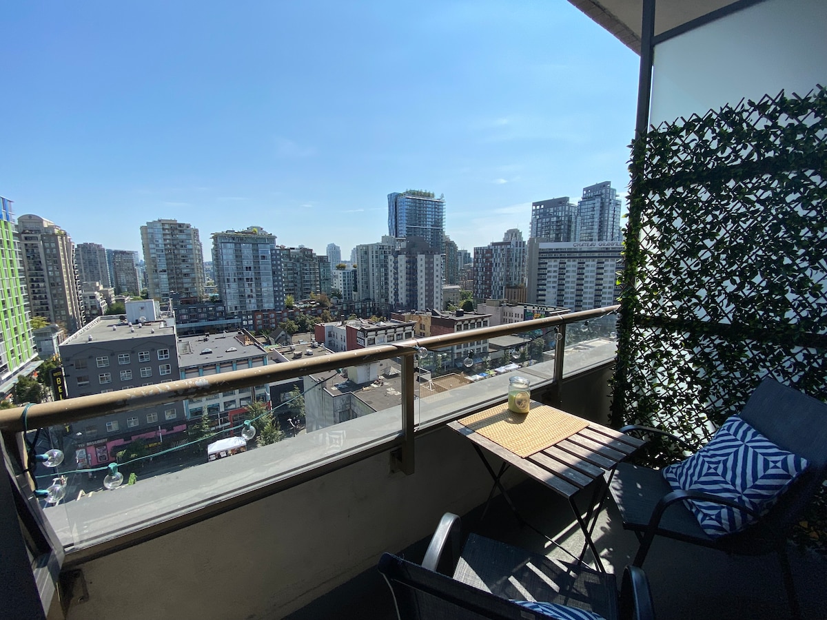 A private balcony overlooks the cityscape, featuring modern high-rise buildings under a clear blue sky. A small table is paired with two chairs, providing a space for relaxation. A decorative plant wall enhances the visual appeal of the balcony.