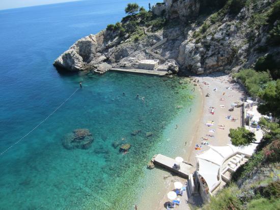 A serene beach view shows clear turquoise waters lapping against a pebbled shoreline. Sunbathers can be seen relaxing on the sand, with rocky cliffs framing the scene in the background. Gentle waves roll in, providing a tranquil atmosphere.