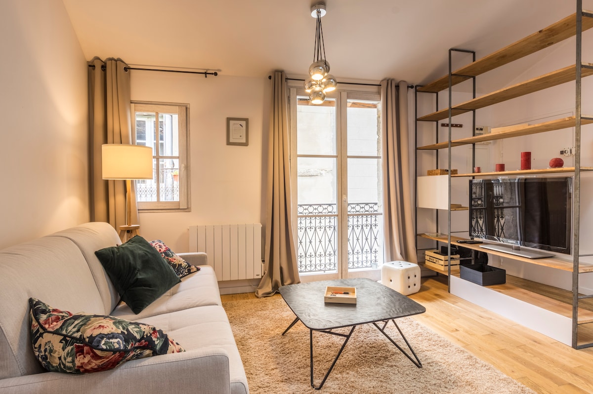 A light-filled living area features a comfortable sofa adorned with decorative cushions. A sleek coffee table is positioned centrally on a plush rug. Two large windows with elegant drapes allow natural light to stream in, while a flat-screen TV rests on a minimalist shelving unit.