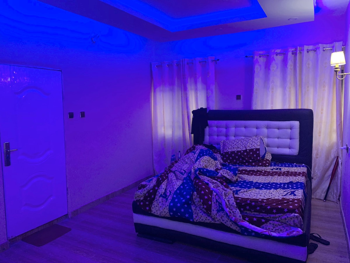 A well-furnished sleeping area features a spacious bed with a patterned quilt. Soft lighting lends a calming, blue hue to the room. Sheer curtains frame the window, allowing natural light to filter in, while a lamp adds additional illumination near the bed.