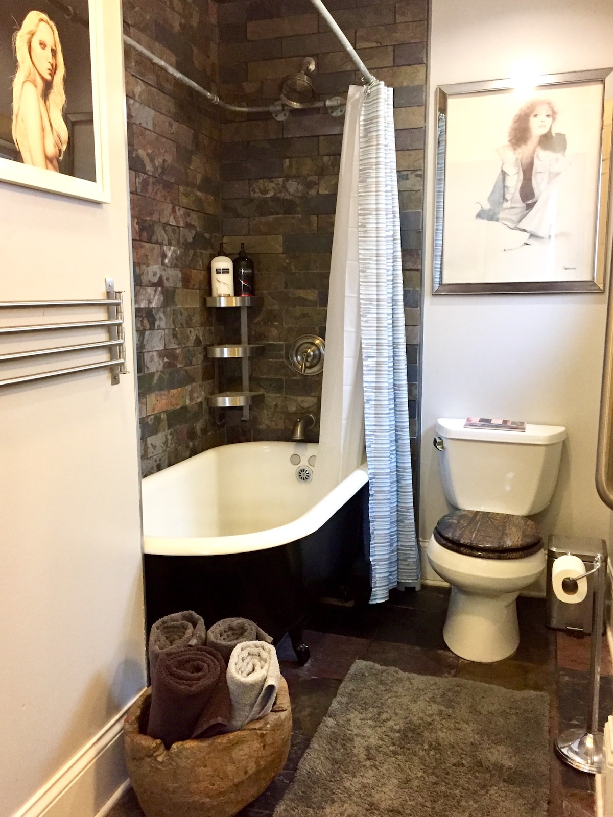 The bathroom features a clawfoot tub accompanied by a shower curtain, and a modern toilet is positioned nearby. Shelving units hold toiletries, while a basket with towels is placed at the tub's foot. Stone wall accents provide texture to the space.