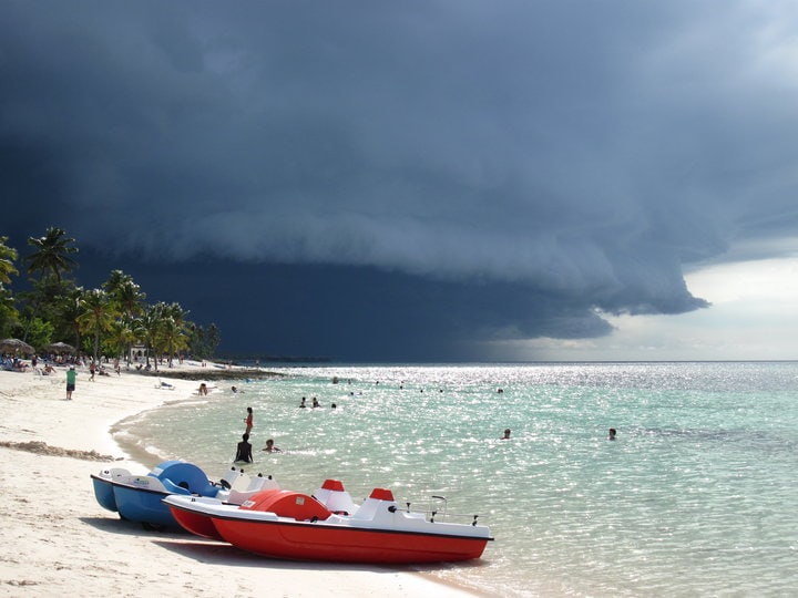 Three pedal boats are lined up on a sandy beach, with their vibrant red and blue colors contrasting against the clear waters. Distinctive dark storm clouds gather in the distance, while beachgoers enjoy the gentle waves under an overcast sky.