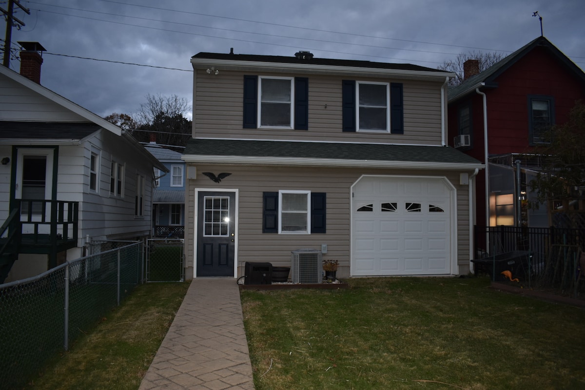 A two-story house is presented, featuring a light grey exterior with dark blue shutters. A front door is highlighted by a small porch, while a garage is integrated on the right. A paved pathway leads toward the entrance, surrounded by a well-kept lawn.
