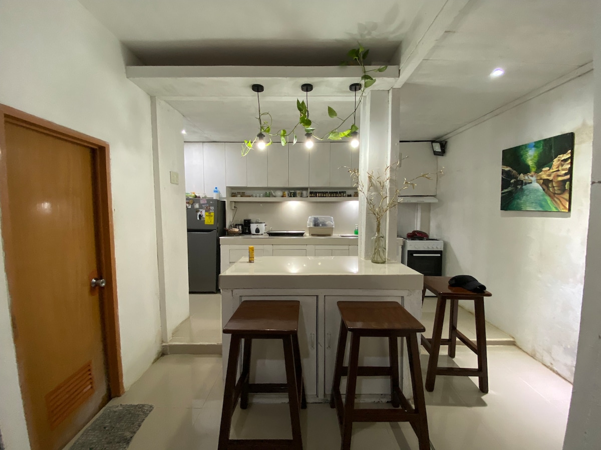 A bright kitchen area features a large island with four wooden stools positioned around it. Minimalistic cabinetry provides ample storage, and modern appliances are visible. Soft overhead lighting illuminates the space, with green plants adding a touch of nature to the decor.