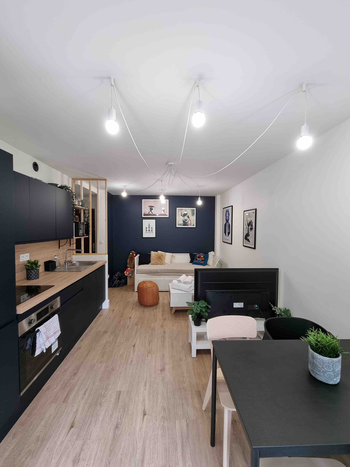 A modern living space is displayed, featuring an open layout. A comfortable seating area includes a sofa and several decorative elements. A dining table is positioned nearby, accompanied by minimalist chairs. The kitchen is equipped with sleek cabinets and appliances, enhancing functionality.