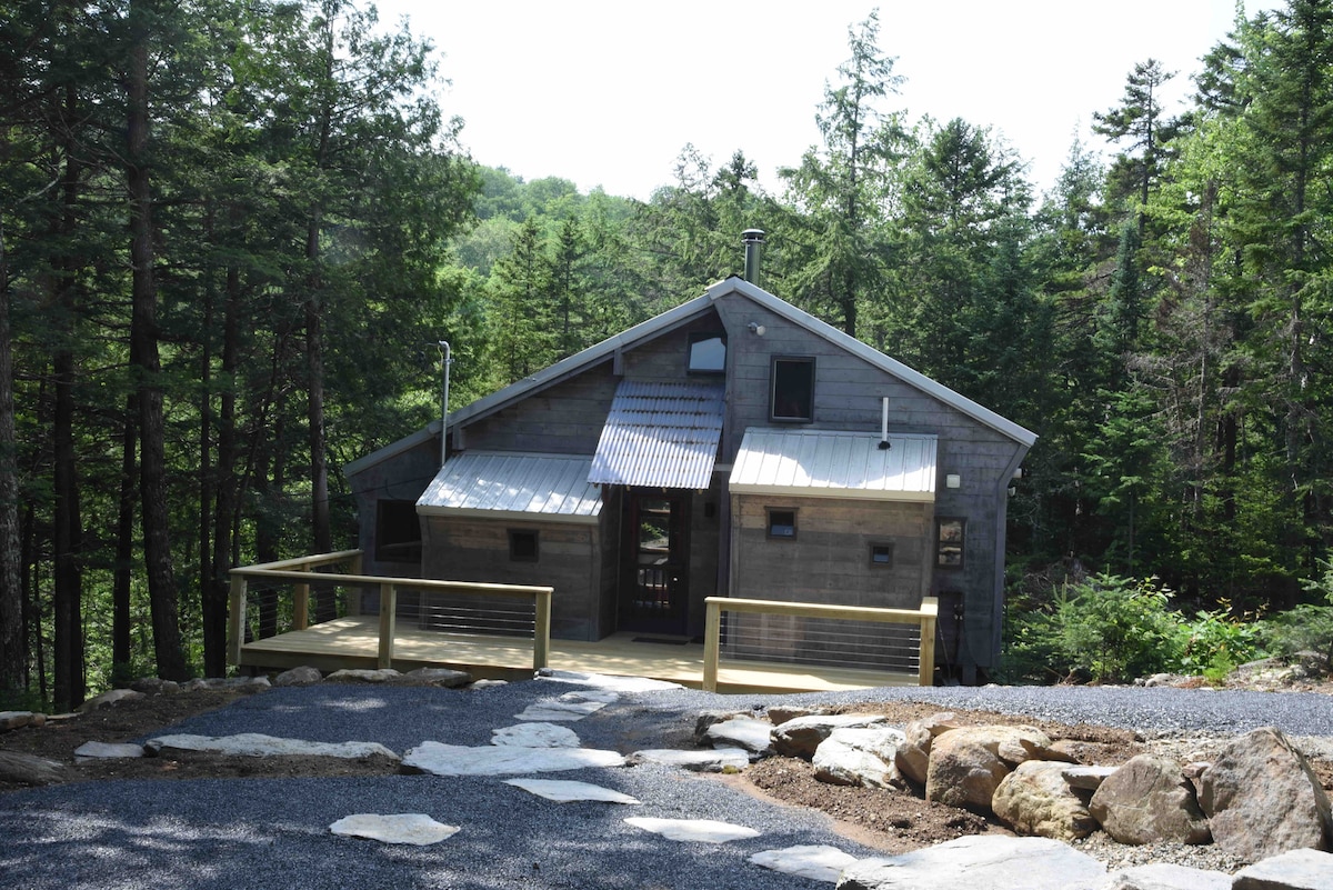 A unique cabin is nestled among lush trees, featuring a wooden deck and a sloped roof. The exterior showcases a combination of natural wood and metal elements, with large windows providing glimpses of the serene forest surroundings.