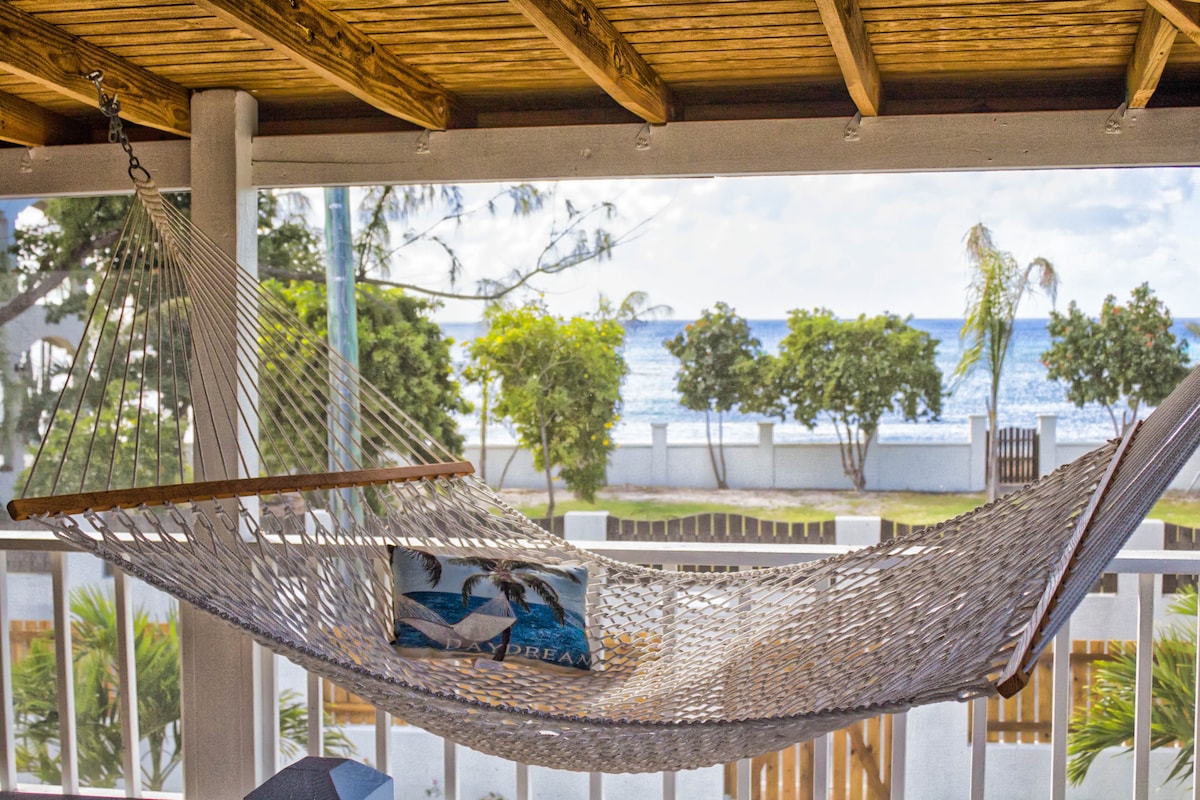 A spacious hammock is suspended on the covered deck, offering views of the ocean in the background. Lush greenery frames the scene, providing a serene atmosphere for relaxation. Natural light pours in, complemented by the wooden beams overhead.