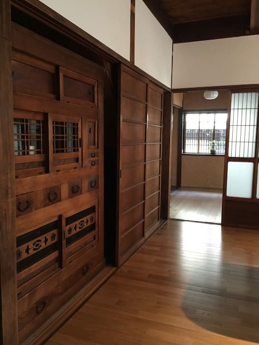 The entrance showcases a traditional wooden door with intricate carvings, leading to a spacious corridor. Light enters through window panels beside sliding doors, illuminating the warm wooden floor. The interior features wooden paneling that complements the historic charm of the house.
