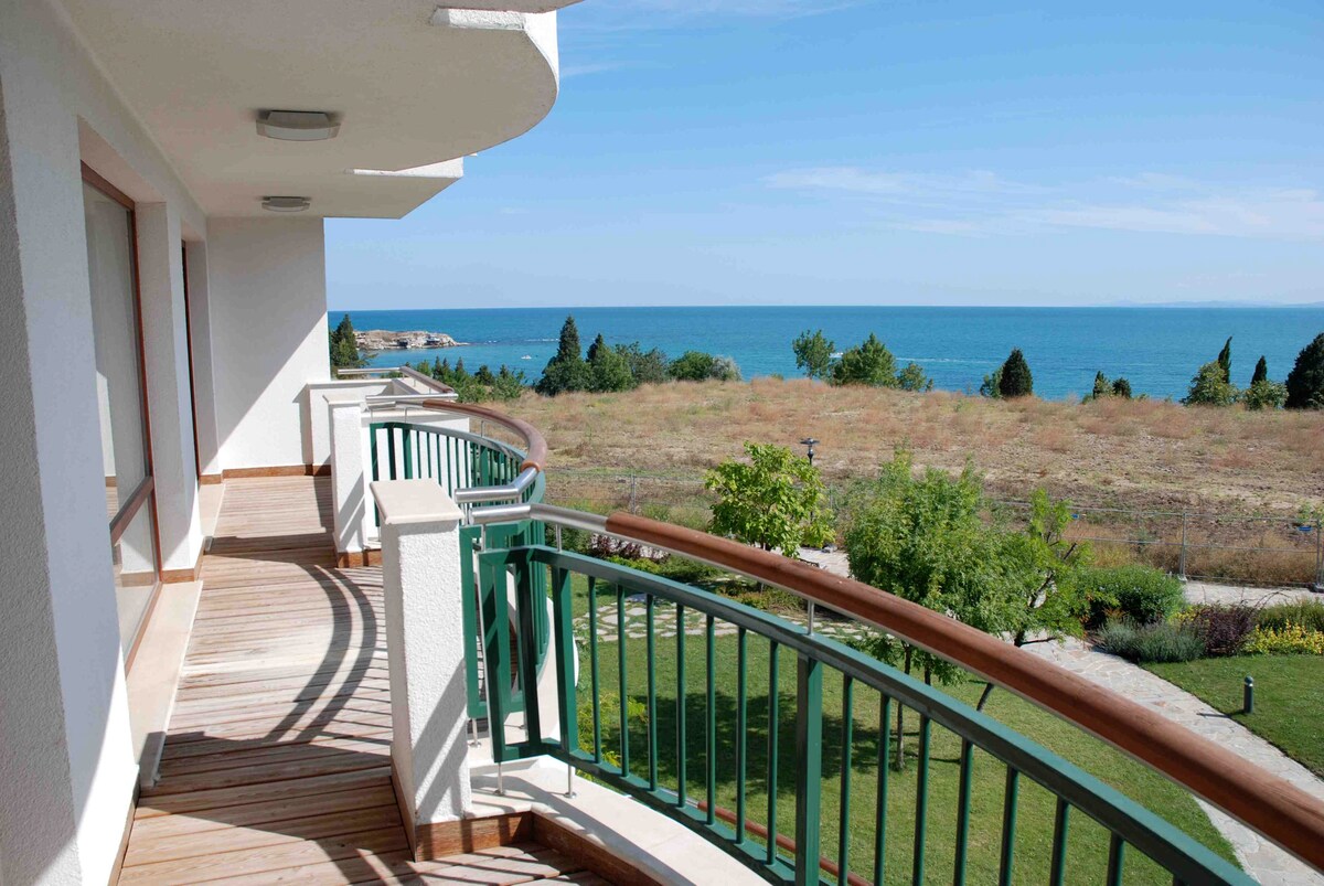 A spacious balcony offers sweeping views of the sea and surrounding greenery. The wooden deck features curved railings and is bathed in natural light, connecting the indoors with the calming outdoor landscape.