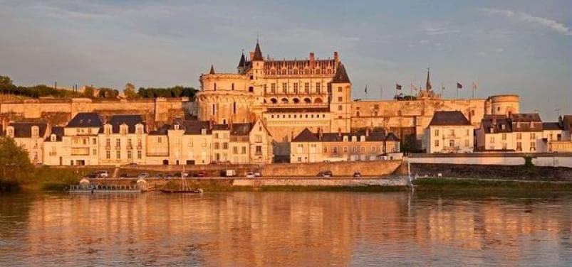 The historic castle is situated atop a hillside, overlooking the calm river. Its elegant facade features turrets and several flagpoles, reflecting golden hues during sunset. Below, a row of quaint buildings lines the water's edge, enhancing the tranquil landscape.
