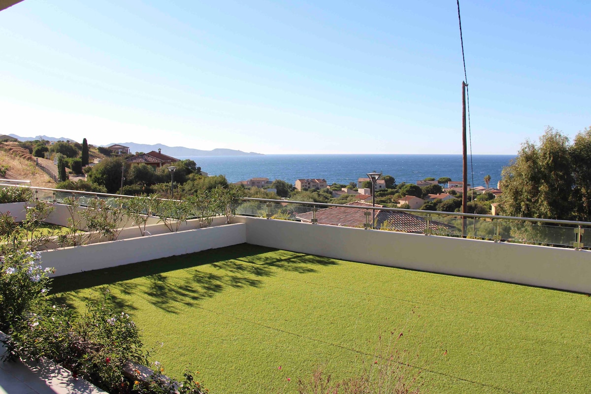 A spacious terrace offers a view of the distant ocean, framed by rolling hills and coastal vegetation. Sunlight illuminates the green lawn, providing an open and inviting space for relaxation. The surrounding area includes distant rooftops and a clear blue sky enhances the tranquil setting.