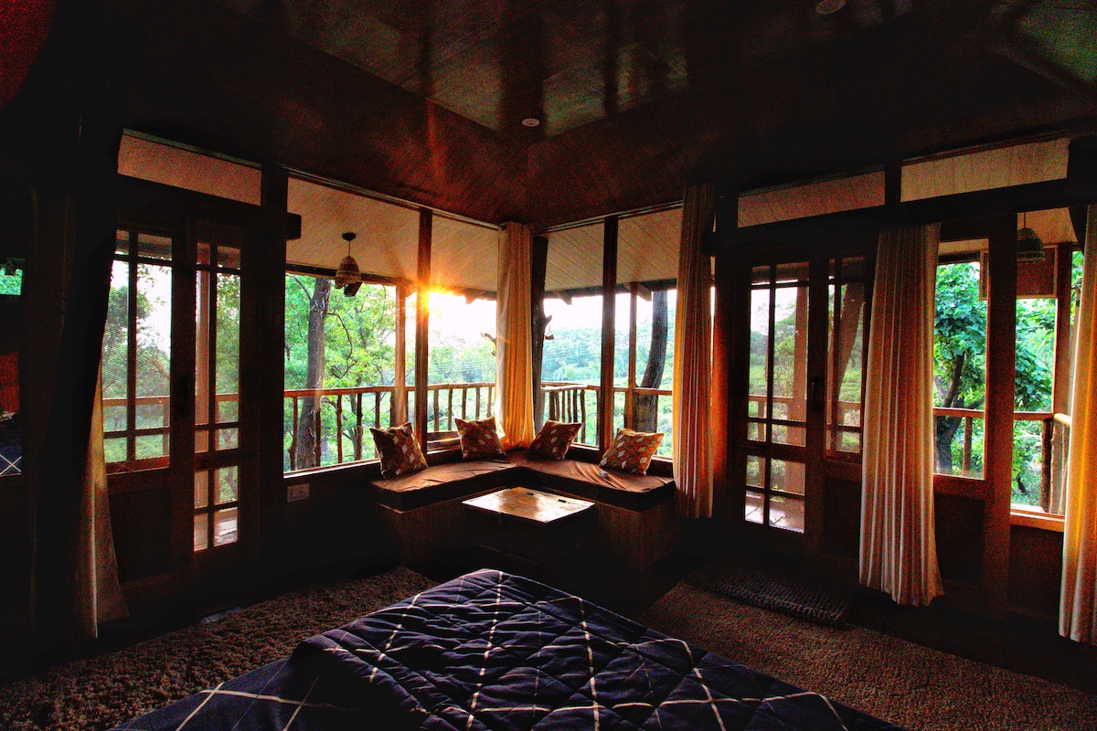 A spacious interior is highlighted by large windows that provide views of the surrounding greenery and sunset. Comfortable seating is arranged along the wall, complemented by soft cushions. Natural light filters through sheer curtains, creating a warm and inviting atmosphere.
