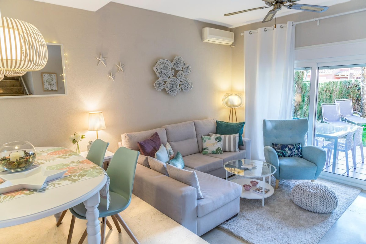 A comfortable living area features a light grey sectional sofa adorned with colorful cushions. A round glass coffee table sits in front of the sofa. A dining space with a floral-patterned table is accompanied by pastel-colored chairs. Natural light enters through sliding doors leading to an outdoor area.