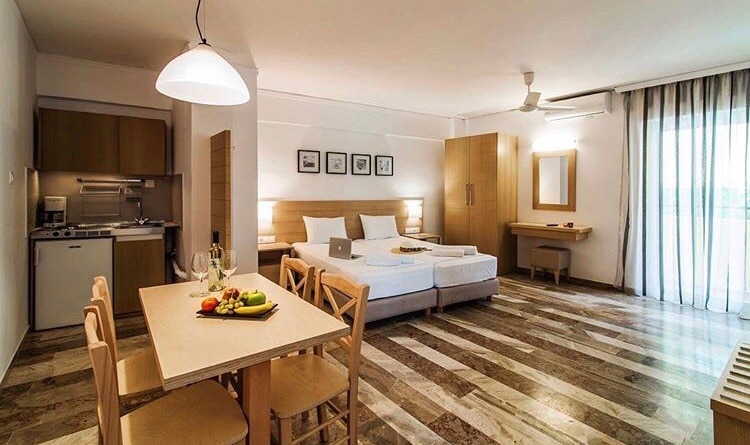 A spacious studio is displayed with a comfortable sleeping area featuring two beds. A dining table with chairs sits in the foreground, accompanied by a fresh fruit arrangement. The kitchenette is visible in the background, and large windows allow natural light to fill the room.
