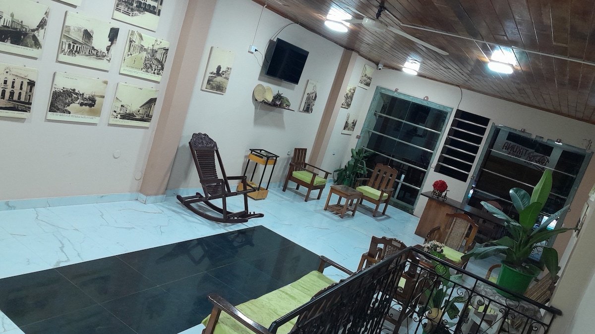An inviting lounge area features a mix of wooden furniture, including rocking chairs and cushioned seats, arranged around a central space. Vintage photographs adorn the walls, while a potted plant adds a touch of greenery. Soft lighting illuminates the room, creating a welcoming atmosphere.