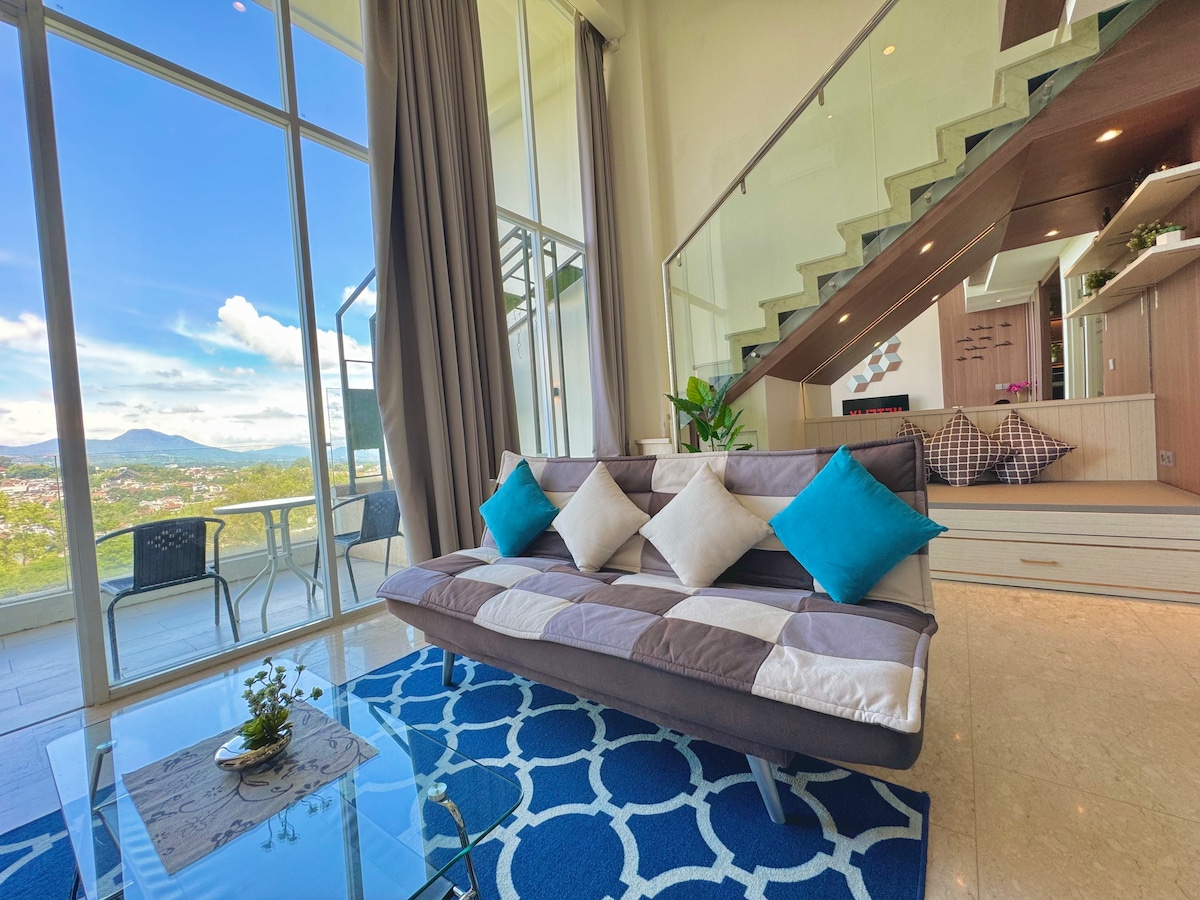A spacious living area features a comfortable sofa adorned with decorative cushions, combining various shades with a grid pattern. Large windows allow natural light to fill the space, showcasing panoramic views of the surrounding landscape. A glass coffee table rests on a patterned area rug, enhancing the elegance of the room.