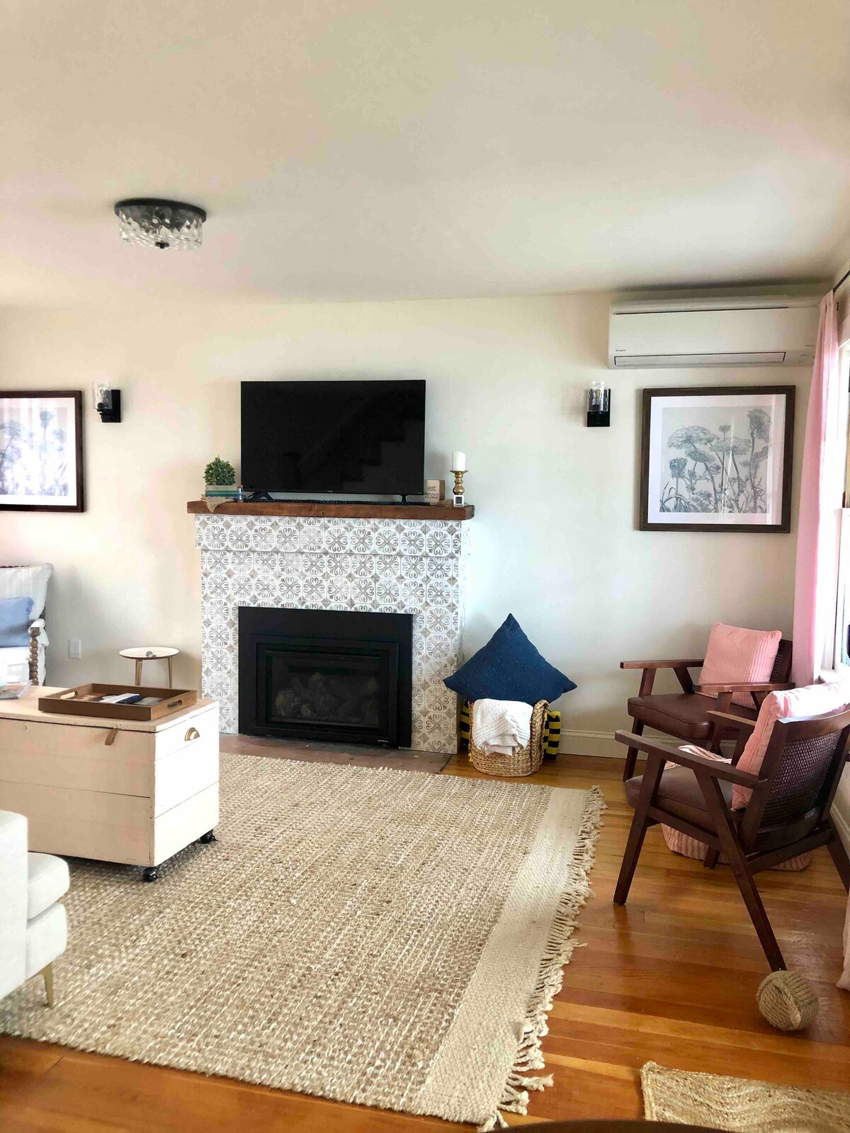 A cozy living area features a styled fireplace with decorative tiles and a mounted television above. A light area rug covers the wooden floor, while comfortable seating options include a white armchair and a wooden chair. Natural light fills the space through nearby windows.