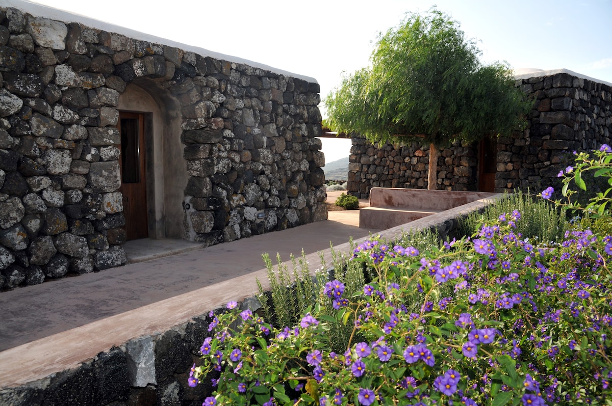 Stone structures are seen nestled among vibrant purple flowers and green foliage, with a pathway leading to the entrance. A tree provides shade in the background, enhancing the natural surroundings.