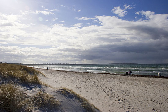 The sandy beach stretches along the coastline, bordered by tall grass on one side. Gentle waves meet the shore under a partly cloudy sky, with varied shades of blue reflected in the water. Small groups of people can be seen enjoying the beach in the distance.