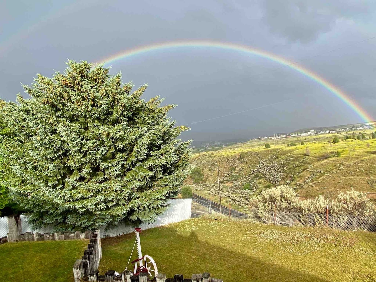 A vibrant rainbow spans across the sky, arching over rolling hills and a grassy area. A large tree can be seen in the foreground, while a few rustic wooden elements are noted near the ground. The landscape hints at a peaceful and scenic outdoor setting.