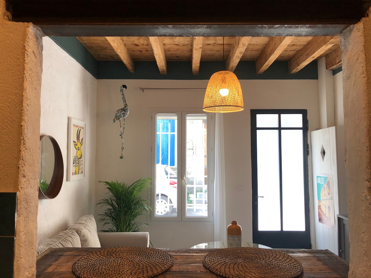 A view from the kitchen area into a bright living space, featuring a cozy sofa and a decorative plant. Large windows allow natural light to fill the room, while a woven pendant light adds a warm touch. A round table with placemats is positioned in the foreground.