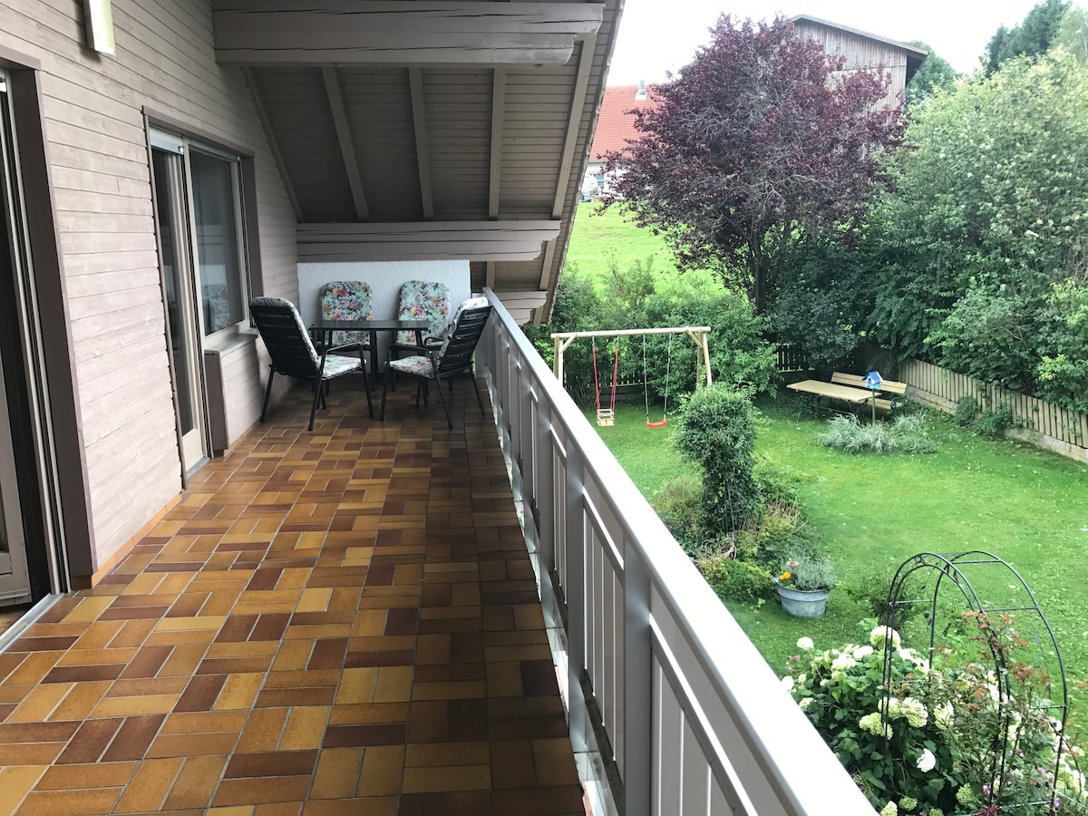A balcony is pictured with a spacious tiled floor. Two chairs and a small table are visible, with a view of a well-kept garden featuring a swing set and greenery. Surrounding trees and hedges provide a natural backdrop.