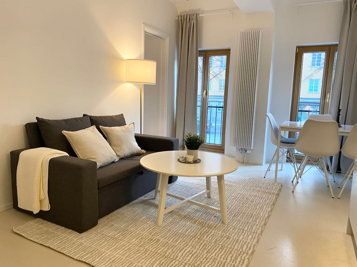 A modern living area is featured, with a gray sofa adorned with light-colored throw pillows. A round table sits in the foreground, accompanied by white chairs. Natural light flows through two windows framed by light curtains, illuminating the neutral color palette and creating a welcoming ambiance.